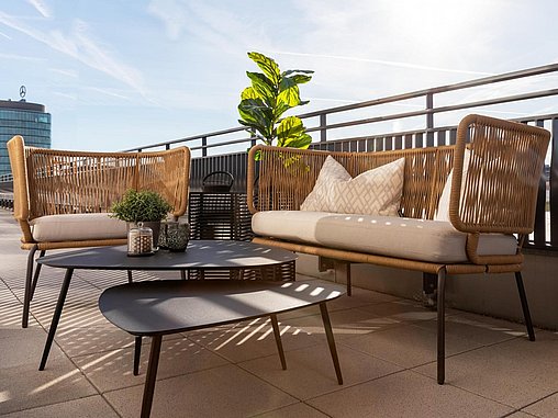 Sonnige Dachterrasse mit gepolsterten Sitzbereich in Rattan-Optik und ovalen Beistelltischen.