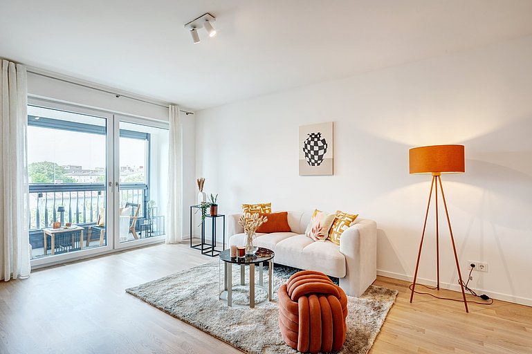 Helles Wohnzimmer mit großen Glasfenstern und Zugang zum Balkon, weißem Sofa mit orangefarbenen und gemusterten Kissen, moderner Stehlampe, rundem Couchtisch und dekorativem Hocker in warmen Farbtönen.