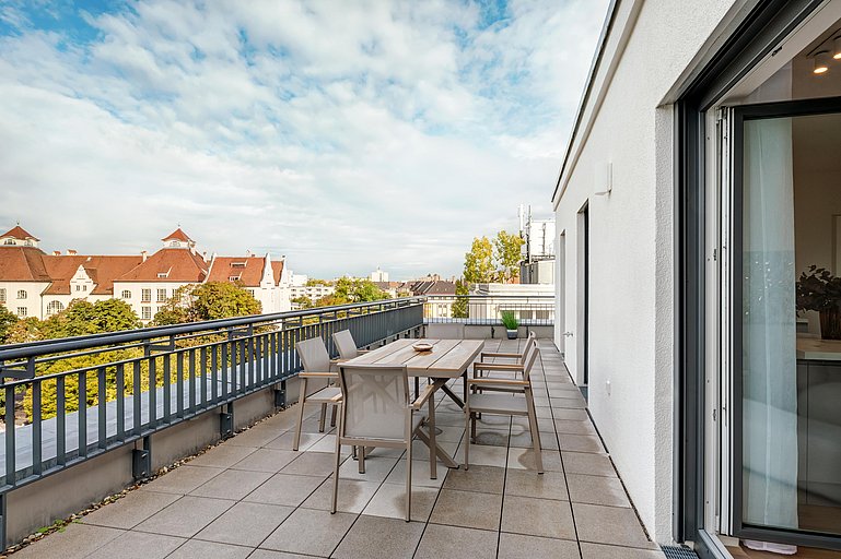 Ein geräumiger Balkon mit Esstisch und Stühlen. Von der Terrasse aus hat man einen Blick auf eine Stadtlandschaft mit Gebäuden mit roten Dächern und Grünflächen unter einem teilweise bewölkten Himmel. Eine offene Glasschiebetür verbindet die Terrasse mit dem Innenbereich.