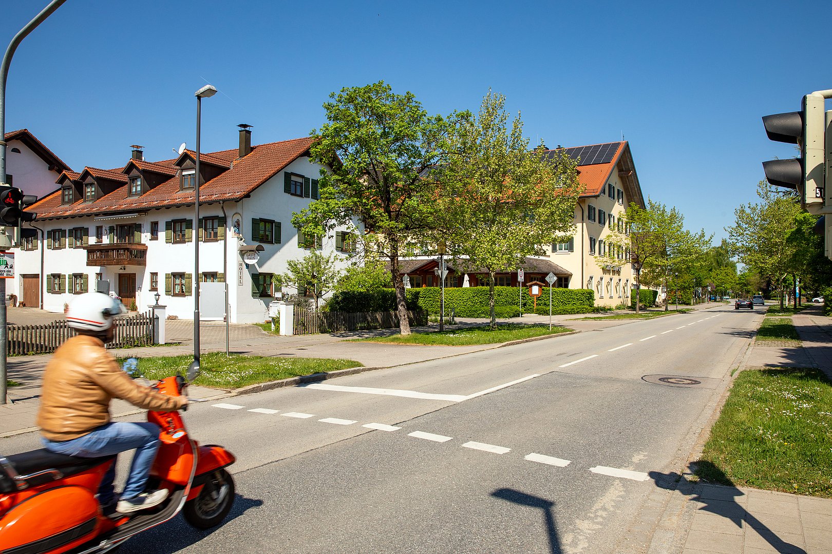 Straßenszene mit Vespafahrer in Aschheim Ortsdurchfahrt in Aschheim mit traditionellen Wohnhäusern, grünen Bäumen und einem Fahrer auf einem roten Motorroller an einer Kreuzung.
