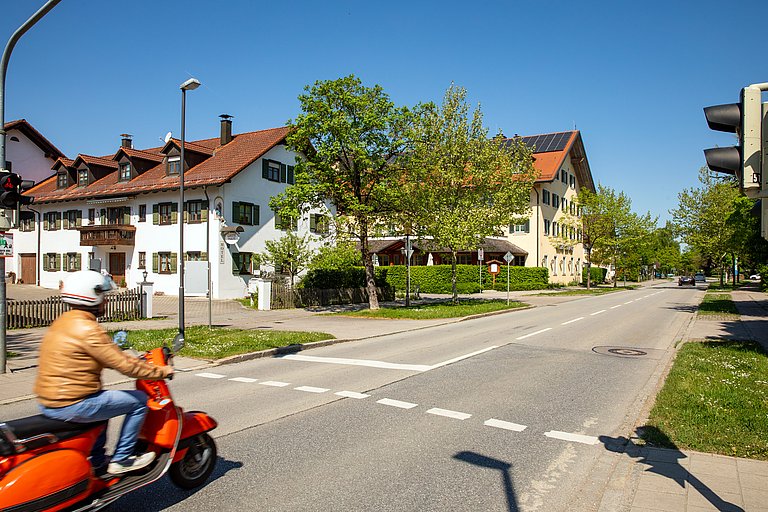Straßenszene mit Vespafahrer in Aschheim Ortsdurchfahrt in Aschheim mit traditionellen Wohnhäusern, grünen Bäumen und einem Fahrer auf einem roten Motorroller an einer Kreuzung.