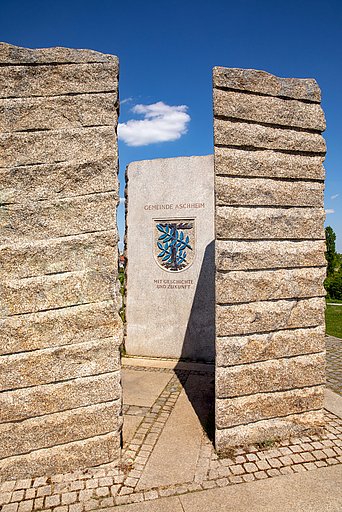 Wappenstein der Gemeinde Aschheim Steinstele der Gemeinde Aschheim mit eingraviertem Schriftzug „Gemeinde Aschheim – Mit Geschichte und Zukunft“ sowie farbigem Gemeindewappen, eingerahmt von zwei massiven Granitpfeilern.