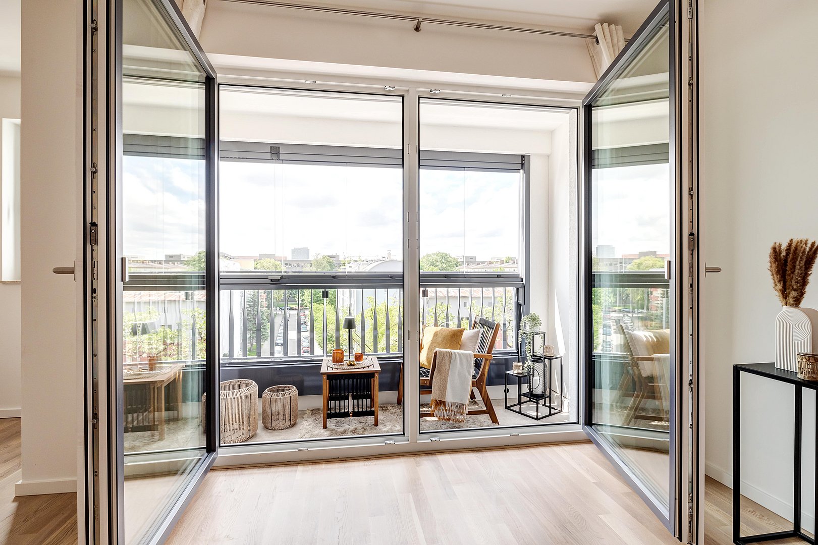 Moderner Balkon mit Glas-Schiebetüren, Boden in Holzoptik, gemütlichem Sitzbereich mit dekorativen Kissen und Decken, kleinem Beistelltisch, stilvollen Körben und Blick auf eine grüne Umgebung.