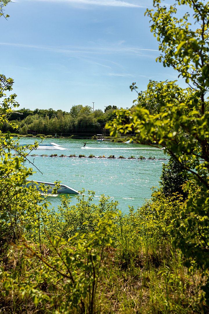 Wasserskianlage in Aschheim Wasserskifahrer auf einer türkisfarbenen Wasserfläche nahe Aschheim bei München, mit Hindernissen im See und umgeben von dichtem Grün an einem sonnigen Tag.