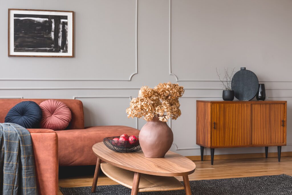 Ein lachsfarbenes Sofa steht vor einer stilvollen Stuckwand. Im Vordergrund ist ein moderner Holztisch zu sehen, auf dem eine bauchige Vase mit einem Trockenblumenstrauß steht. Die Vintage-Kommode im Hintergrund sorgt für einen modernen Stilbruch.