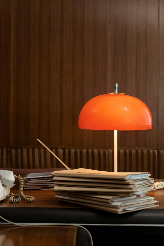 Eine Lampe im Retro-Design steht auf einem Schreibtisch. Die Lampe hat einen orangefarbenen Lampenschirm und ist in typischer Pilzform gestaltet. Vor der Lampe liegt ein Heftstapel, und links davon steht ein Telefon. Die hölzerne Wand im Hintergrund harmoniert mit dem Vintage-Design des Zimmers.